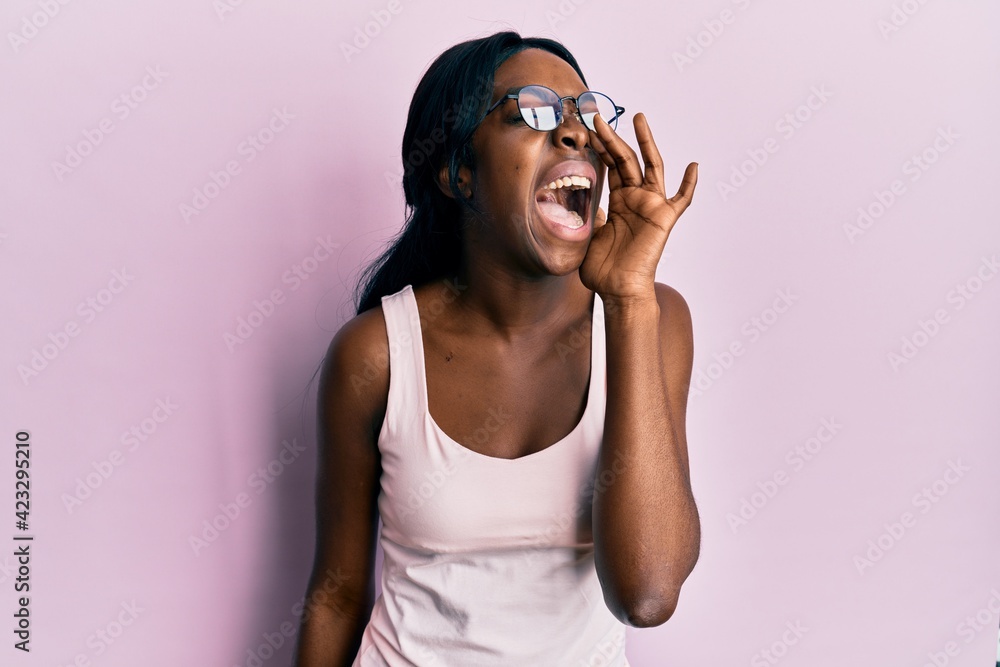 Young african american woman wearing casual clothes and glasses shouting and screaming loud to side with hand on mouth. communication concept. Young african american woman wearing casual clothes and glasses shouting and screaming loud to side with hand on mouth. communication concept.