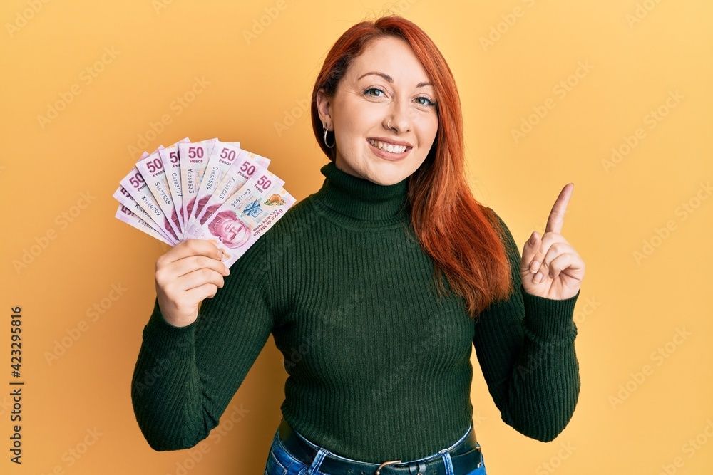Beautiful redhead woman holding 50 mexican pesos banknotes smiling happy pointing with hand and finger to the side Beautiful redhead woman holding 50 mexican pesos banknotes smiling happy pointing with hand and finger to the side