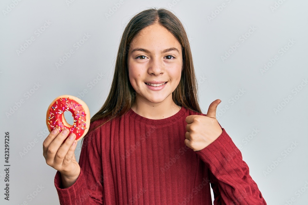 Beautiful brunette little girl holding tasty colorful doughnut smiling happy and positive, thumb up doing excellent and approval sign Beautiful brunette little girl holding tasty colorful doughnut smiling happy and positive, thumb up doing excellent and approval sign
