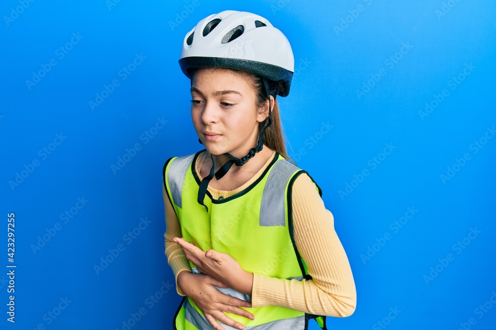 Beautiful brunette little girl wearing bike helmet and reflective vest with hand on stomach because nausea, painful disease feeling unwell. ache concept. Beautiful brunette little girl wearing bike helmet and reflective vest with hand on stomach because nausea, painful disease feeling unwell. ache concept.