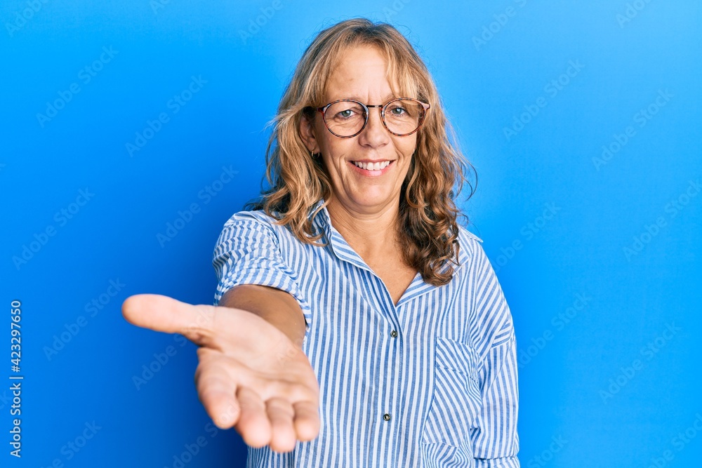 Middle age blonde woman wearing casual clothes and glasses smiling friendly offering handshake as greeting and welcoming. successful business. Middle age blonde woman wearing casual clothes and glasses smiling friendly offering handshake as greeting and welcoming. successful business.