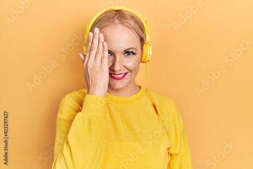 Young blonde woman listening to music using headphones covering one eye with hand, confident smile on face and surprise emotion.