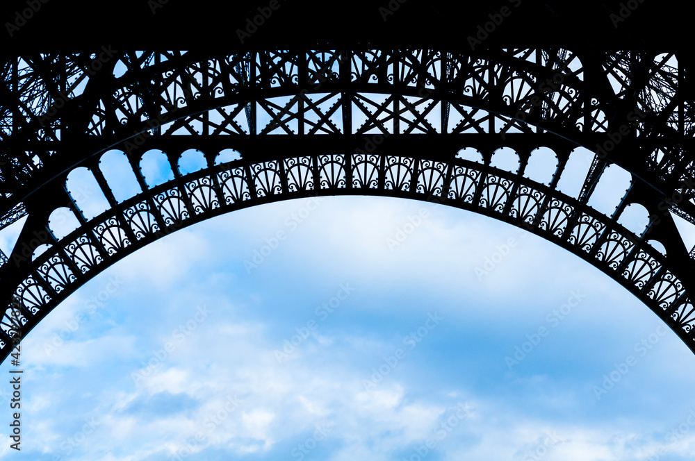 Arched detail of the Eiffel Tower silhouetted against a cloudy blue sky.