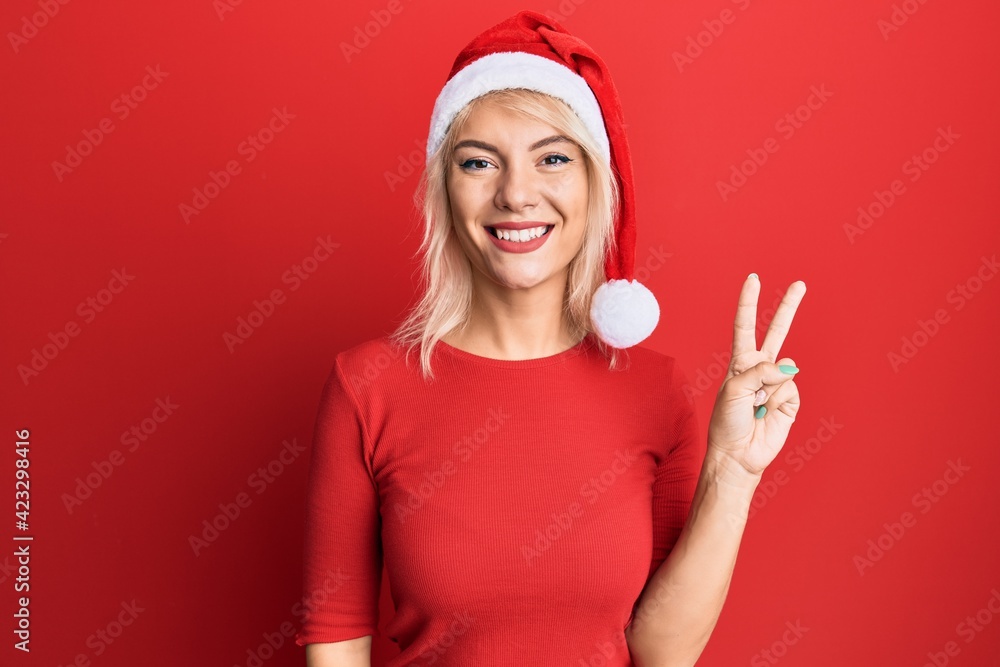 Young blonde girl wearing christmas hat smiling with happy face winking at the camera doing victory sign. number two. Young blonde girl wearing christmas hat smiling with happy face winking at the camera doing victory sign. number two.