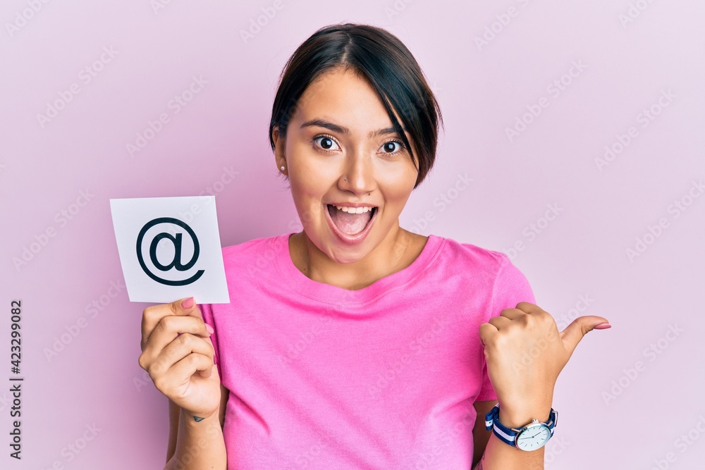 Beautiful young woman with short hair holding paper with email symbol pointing thumb up to the side smiling happy with open mouth Beautiful young woman with short hair holding paper with email symbol pointing thumb up to the side smiling happy with open mouth