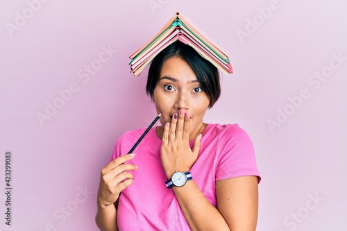 Beautiful young woman with short hair holding book over head covering mouth with hand, shocked and afraid for mistake. surprised expression