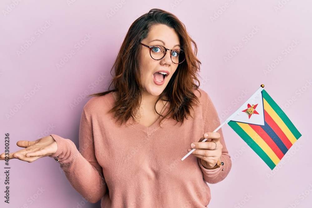 Young caucasian woman holding zimbabwe flag celebrating achievement with happy smile and winner expression with raised hand Young caucasian woman holding zimbabwe flag celebrating achievement with happy smile and winner expression with raised hand