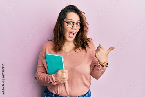 Young caucasian woman holding book pointing thumb up to the side smiling happy with open mouth