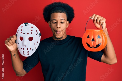 African american man with afro hair wearing hockey mask and halloween pumpking making fish face with mouth and squinting eyes, crazy and comical. photo