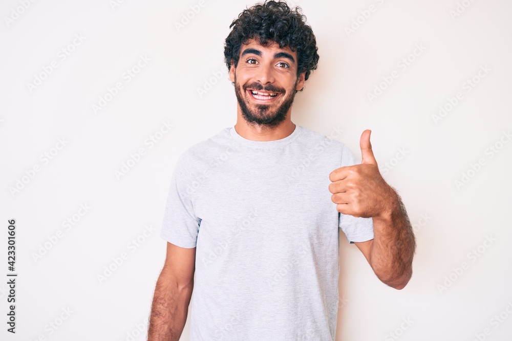Handsome young man with curly hair and bear wearing casual tshirt smiling happy and positive, thumb up doing excellent and approval sign Handsome young man with curly hair and bear wearing casual tshirt smiling happy and positive, thumb up doing excellent and approval sign