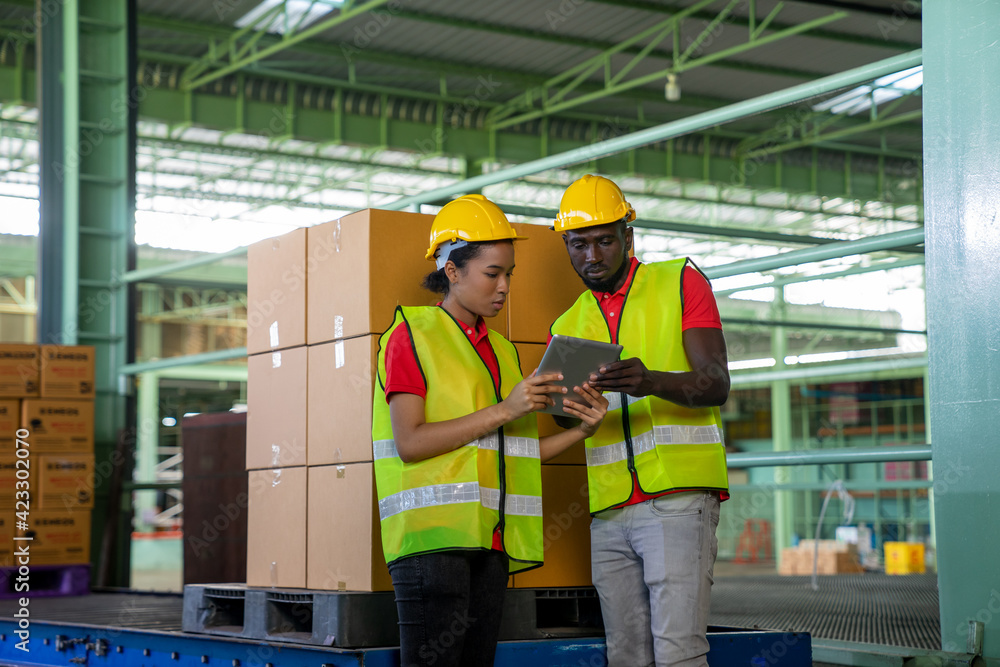 African American dispatchers use tablets to checking delivery order for dispatch in industrial warehouse. Black female worker and male carrier in safety vest, hard hat working in transport service. African American dispatchers use tablets to checking delivery order for dispatch in industrial warehouse. Black female worker and male carrier in safety vest, hard hat working in transport service.