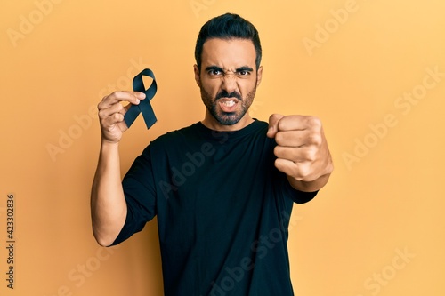 Young hispanic man holding black ribbon annoyed and frustrated shouting with anger, yelling crazy with anger and hand raised