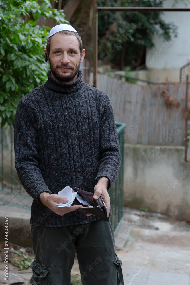 Obraz premium Man showing empty wallet. Jewish man wearing kippah in Hebrew or yarmulke with green trees in the background, beard wearing black sweatshirt. No money concept against the background of the pandemic