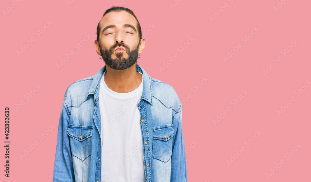 Fototapeta premium Attractive man with long hair and beard wearing casual denim jacket looking at the camera blowing a kiss on air being lovely and sexy. love expression.
