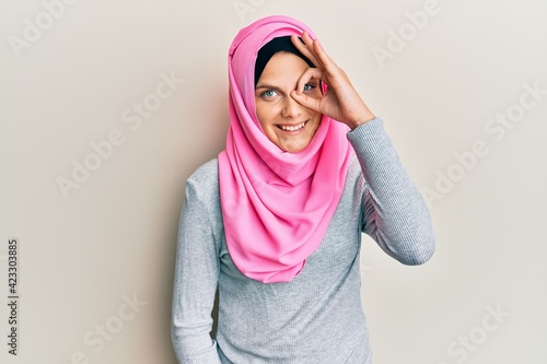 Young caucasian woman wearing traditional islamic hijab scarf doing ok gesture with hand smiling, eye looking through fingers with happy face. © Krakenimages.com