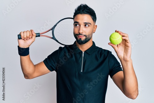 Young man with beard playing tennis holding racket and ball smiling looking to the side and staring away thinking. © Krakenimages.com