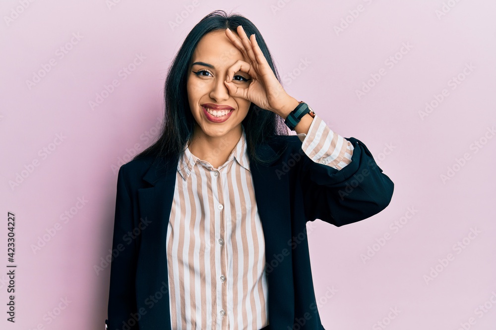 Young hispanic girl wearing business clothes doing ok gesture with hand smiling, eye looking through fingers with happy face. Young hispanic girl wearing business clothes doing ok gesture with hand smiling, eye looking through fingers with happy face.