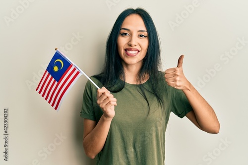 Young hispanic girl holding malaysia flag smiling happy and positive, thumb up doing excellent and approval sign