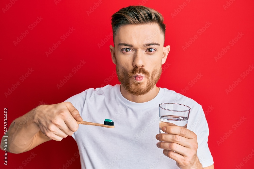 Young redhead man holding toothbrush with toothpaste and glass of water making fish face with mouth and squinting eyes, crazy and comical. Young redhead man holding toothbrush with toothpaste and glass of water making fish face with mouth and squinting eyes, crazy and comical.