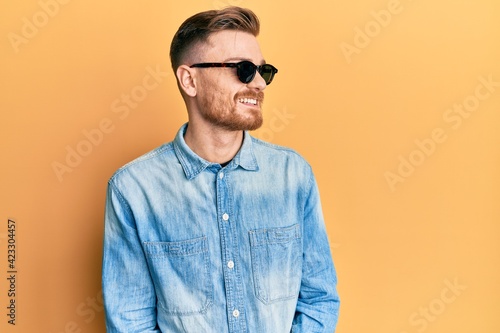 Young redhead man wearing stylish sunglasses looking away to side with smile on face, natural expression. laughing confident.