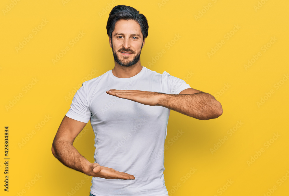 Young hispanic man wearing casual white t shirt gesturing with hands showing big and large size sign, measure symbol. smiling looking at the camera. measuring concept. Young hispanic man wearing casual white t shirt gesturing with hands showing big and large size sign, measure symbol. smiling looking at the camera. measuring concept.