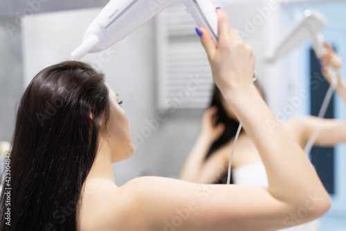 Beautiful woman using a hair dryer and smiling while looking at the mirror. Smiling woman drying hair with hair dry machine. Happy woman looking at mirror while using hair dryer in the bathroom.