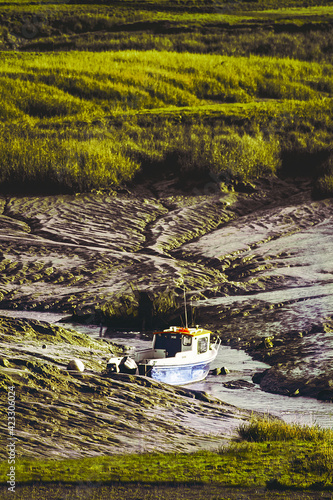 A blue and red boat moored to the shore of a dry river. The boat got stuck in the mud. Artistic patterns on the ground. In the distance, a green grassy meadow.