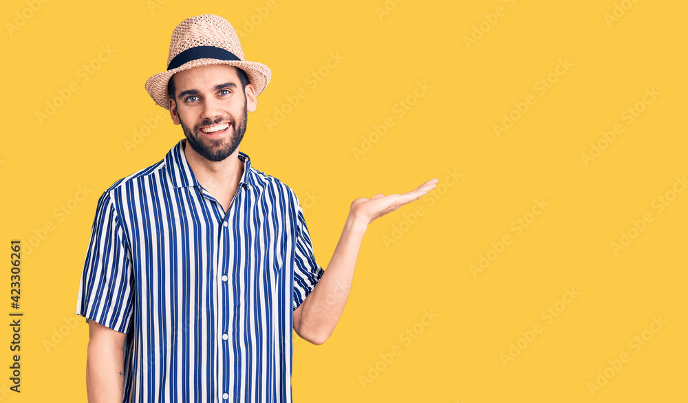 Young handsome man with beard wearing summer hat and striped shirt smiling cheerful presenting and pointing with palm of hand looking at the camera.