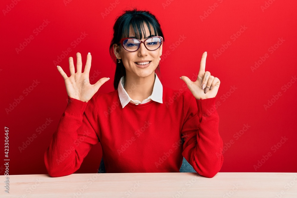 Young brunette woman with bangs wearing glasses sitting on the table showing and pointing up with fingers number seven while smiling confident and happy. Young brunette woman with bangs wearing glasses sitting on the table showing and pointing up with fingers number seven while smiling confident and happy.