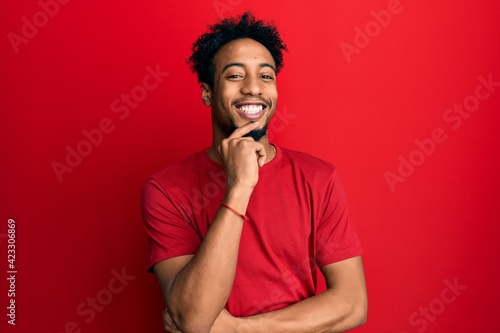 Young african american man with beard wearing casual red t shirt looking confident at the camera with smile with crossed arms and hand raised on chin. thinking positive. © Krakenimages.com