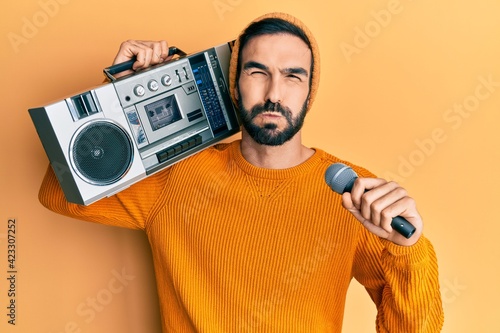 Young hispanic man holding boombox, listening to music singing with microphone puffing cheeks with funny face. mouth inflated with air, catching air.