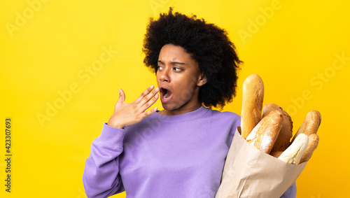 Young African American woman buying something bread isolated on yellow background yawning and covering wide open mouth with hand