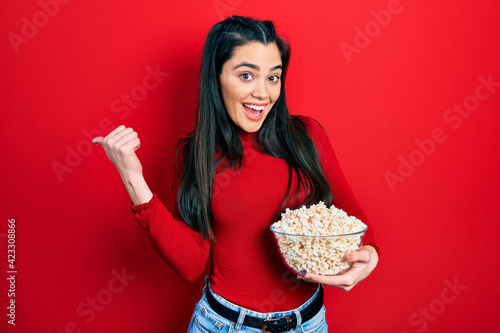 Young hispanic girl eating popcorn pointing thumb up to the side smiling happy with open mouth