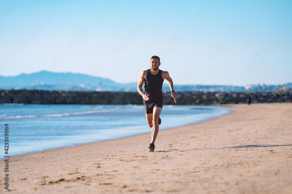 Runner in motion. Young sporty athletic man in black sportswear running or jogging along the beach near the ocean. Healthy lifestyle