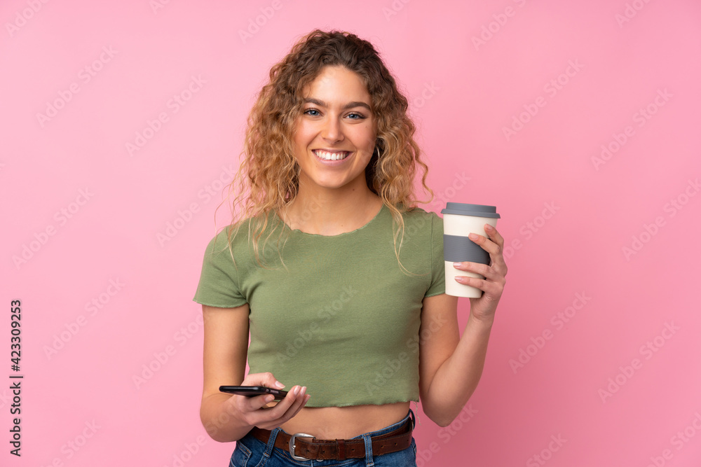 Young blonde woman with curly hair isolated on pink background holding coffee to take away and a mobile Young blonde woman with curly hair isolated on pink background holding coffee to take away and a mobile