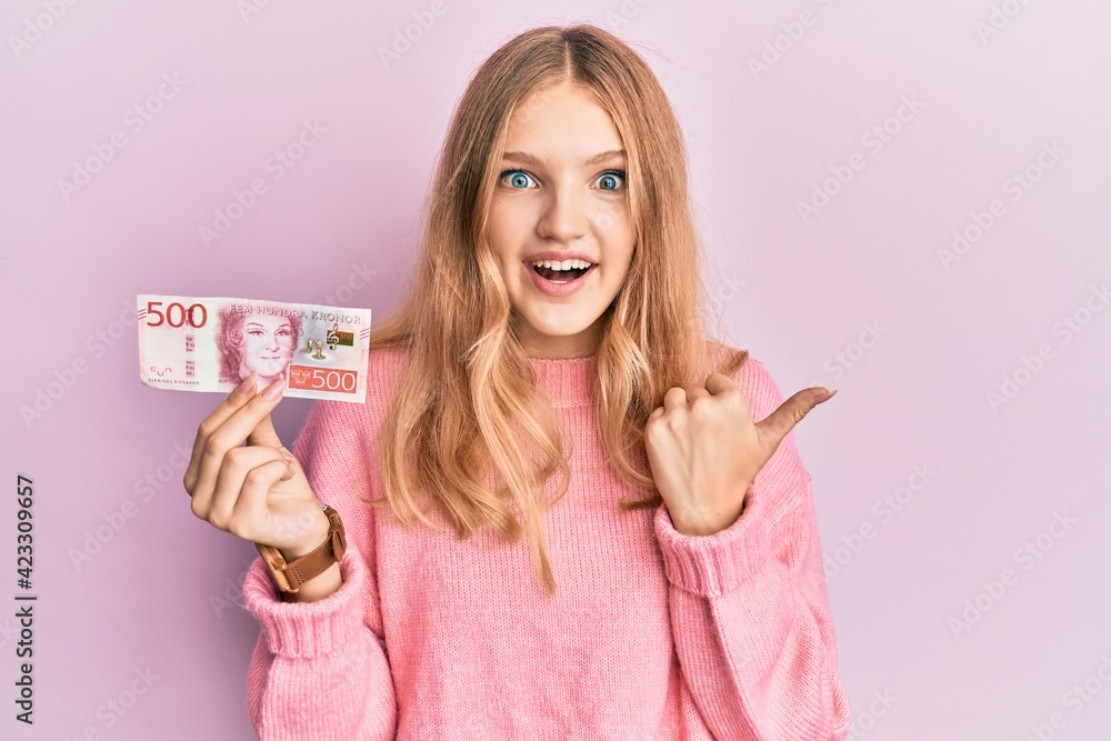 Beautiful young caucasian girl holding 500 swedish krona banknote pointing thumb up to the side smiling happy with open mouth Beautiful young caucasian girl holding 500 swedish krona banknote pointing thumb up to the side smiling happy with open mouth