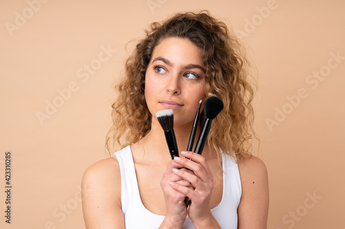 Young blonde woman with curly hair isolated on beige background holding makeup brush and looking up