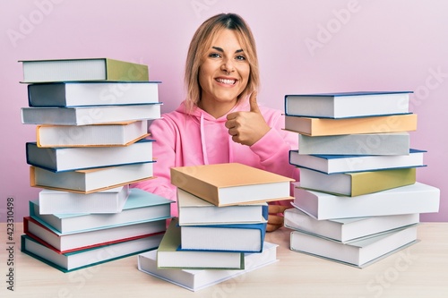 Young caucasian woman sitting on the table with books doing happy thumbs up gesture with hand. approving expression looking at the camera showing success.