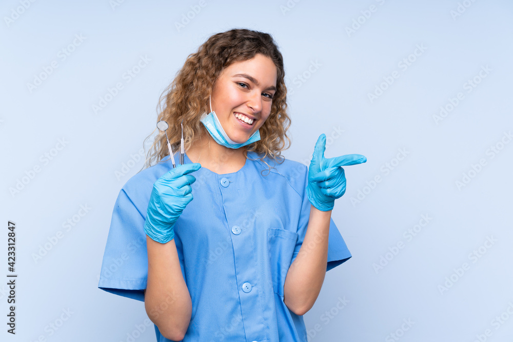 Young blonde woman dentist holding tools isolated on blue background pointing finger to the side Young blonde woman dentist holding tools isolated on blue background pointing finger to the side