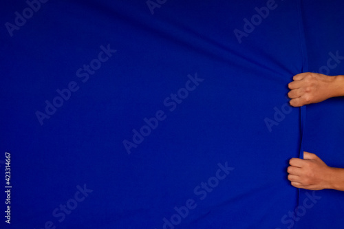 pair of a man's hands grasping a curtain and clenching his fists, as if he were lifting the blue fabric or dragging it, blue background with plenty of room for text