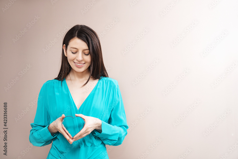 Brunette young woman holding heart in hands. Plain wall background. Wearing light blue dress. Copy space for text. Ad concept Brunette young woman holding heart in hands. Plain wall background. Wearing light blue dress. Copy space for text. Ad concept