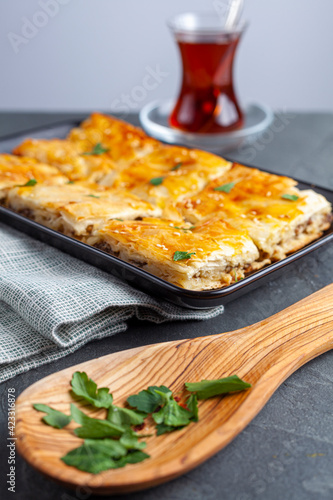 Turkish kiymali borek served on a tray after being sliced into square shapes. Fresh parsley leaves are sprinkled on top for added flavor. A glass of Turkish tea in special tea glass nearby. photo