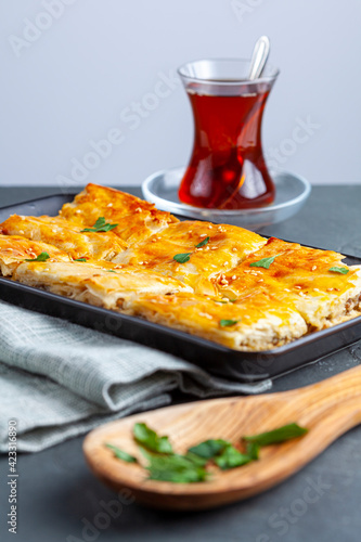 Turkish kiymali borek served on a tray after being sliced into square shapes. Fresh parsley leaves are sprinkled on top for added flavor. A glass of Turkish tea in special tea glass nearby. photo