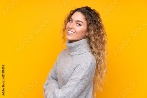 Young blonde woman with curly hair wearing a turtleneck sweater isolated on yellow background with arms crossed and looking forward © luismolinero