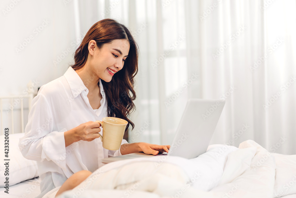 Young smiling happy beautiful asian woman relaxing using laptop computer and drinking coffee in the bedroom at home. Young creative girl working and typing on keyboard.work from home concept Young smiling happy beautiful asian woman relaxing using laptop computer and drinking coffee in the bedroom at home. Young creative girl working and typing on keyboard.work from home concept