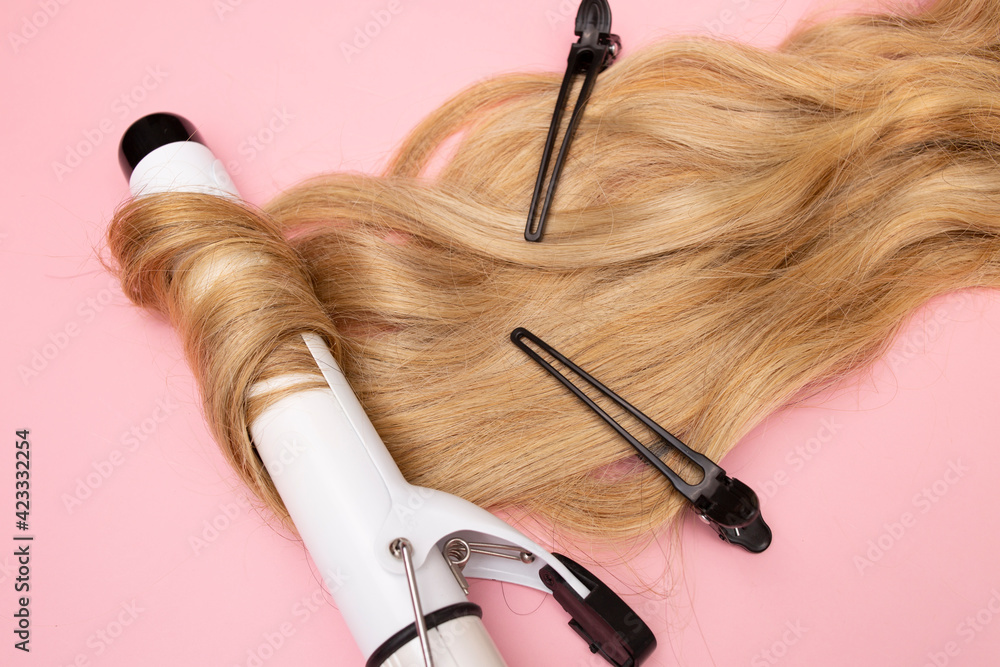Naklejka premium Curling blonde hair on a large diameter curling iron on a pink background. Curl care, hair styling, black clips.