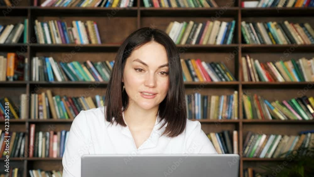Woman freelancer with short hair talks and nods at online job interview near grey laptop against large bookshelves in local library zoom in Woman freelancer with short hair talks and nods at online job interview near grey laptop against large bookshelves in local library zoom in
