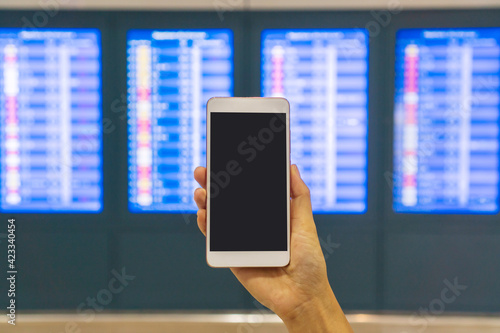 Hand of young Asian tourist woman holding mobile phone near flight information board in international airport. Close up, copy space