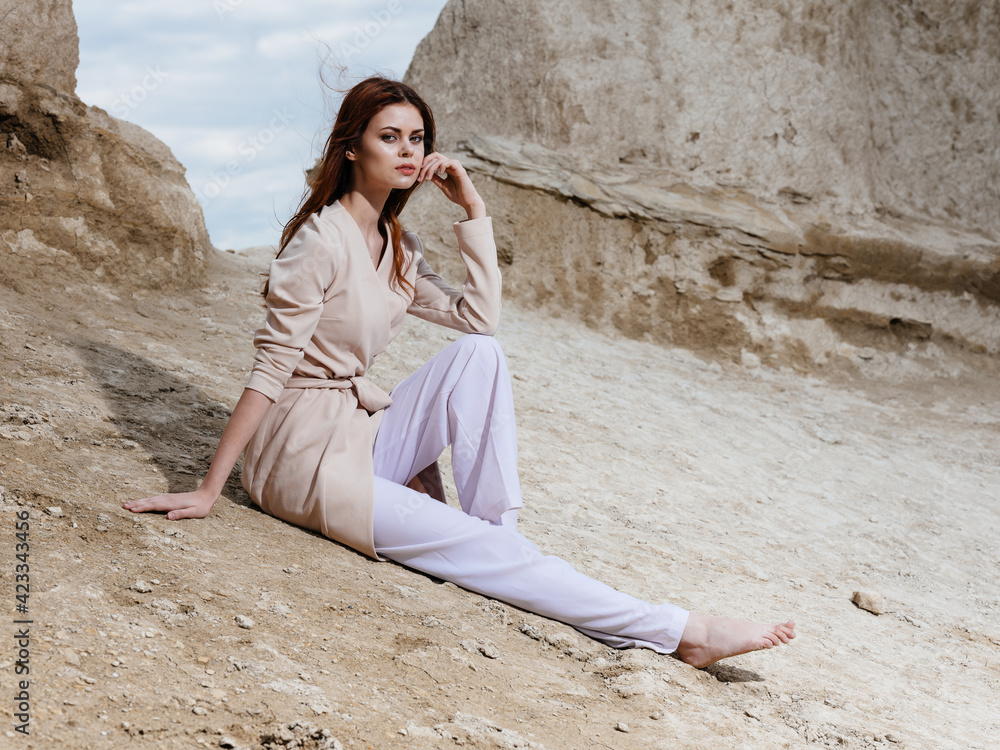 woman in light clothes posing sitting on the sand near the rocks woman in light clothes posing sitting on the sand near the rocks