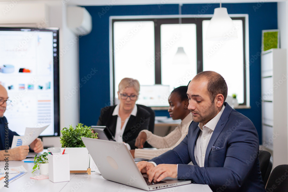 Busy business man using laptop typing sitting at conference table in broadroom concentrated on job. Diverse colleagues working in background. Multiethnic coworkers planning new financial project. Busy business man using laptop typing sitting at conference table in broadroom concentrated on job. Diverse colleagues working in background. Multiethnic coworkers planning new financial project.